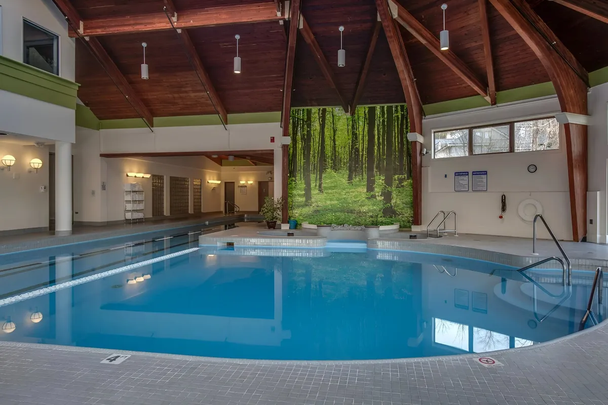 An indoor swimming pool scene featuring a wooden ceiling and a calming forest mural, adding to the serene, spa-like atmosphere. The pool area is spacious and designed for both individual relaxation and social gatherings.