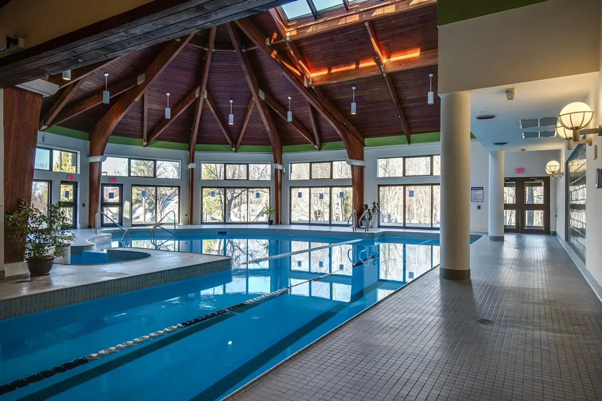 A bright, modern indoor swimming pool with large windows allowing sunlight to stream in, a wooden ceiling, and skylights. The space exudes a calming, rejuvenating atmosphere, ideal for relaxing swims or casual socializing by the water.