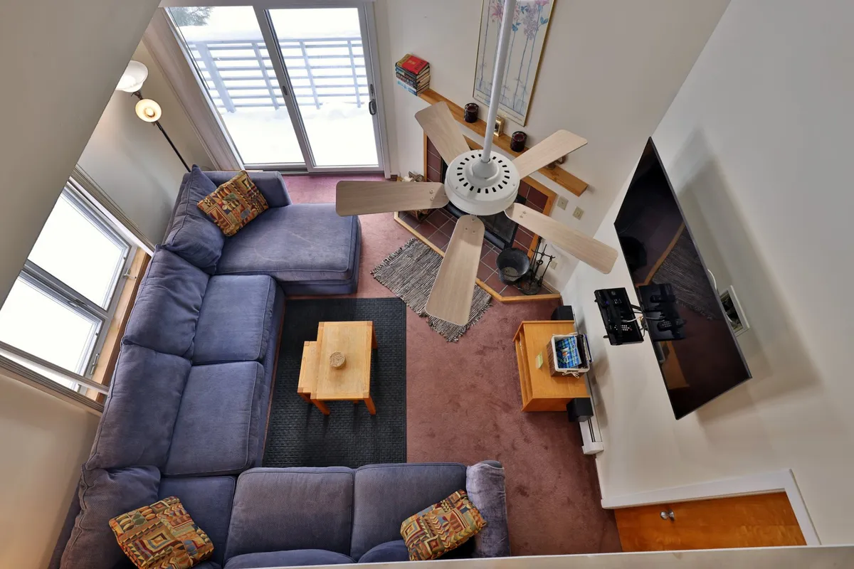 A spacious living area with a large sectional sofa, wooden coffee table, ceiling fan, television, and sliding glass door. The room is designed for both relaxation and socializing, offering additional sleeping space via the sleeper sofa.