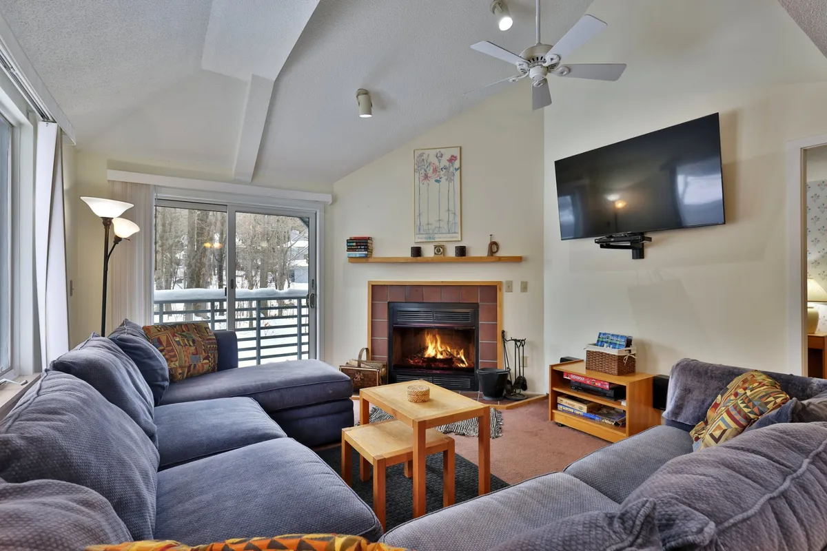 The image features a warm, inviting living area centered around a crackling fireplace, with a plush sofa and coffee table set for gathering. A television is positioned for cozy movie nights, and the overall scene radiates comfort and togetherness, perfect for après-ski relaxation or unwinding after a day exploring Killington.