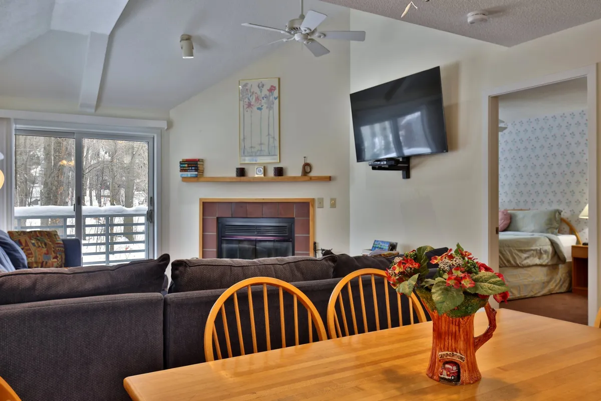 A spacious, open-plan living room and dining area featuring a large sectional sofa, a cozy wood-burning fireplace, a dining table, television, and tasteful wall art. Large windows let in ample natural light and offer glimpses of the outdoors, while the arrangement encourages both relaxation and communal gatherings.