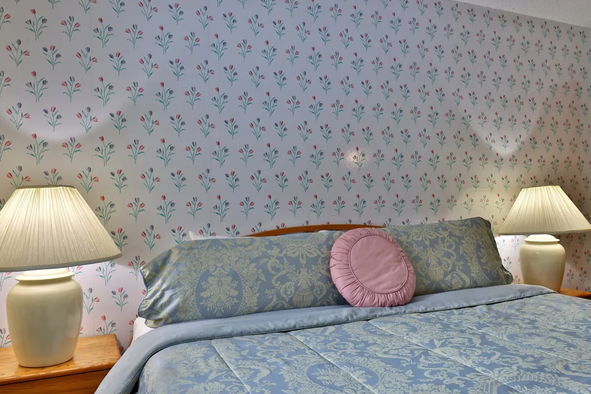 A tranquil bedroom scene with a comfortable bed adorned with decorative pillows, soft lighting from bedside lamps, and floral patterned wallpaper. The overall mood is one of calm and serenity, ideal for restful nights.