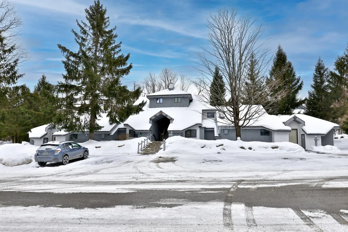An exterior shot of the property featuring a snow-covered ground, tall evergreen trees, the building's architectural design, and a driveway leading up to the entrance. The scene suggests spacious grounds and a tranquil natural setting, though in winter.