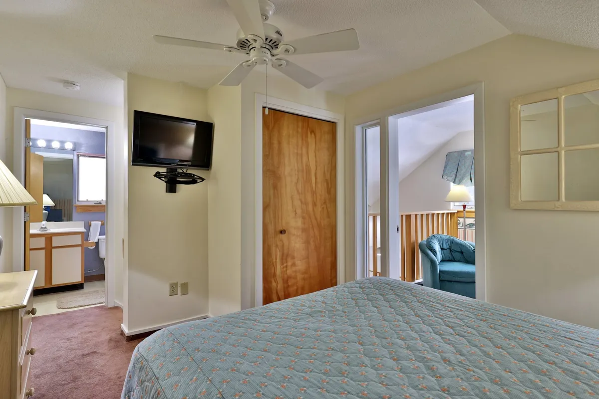 A welcoming bedroom with a quilted bed, TV mounted on the wall, a door leading to another room, and a light blue chair. The space exudes warmth, providing a private sanctuary for relaxation or enjoying a movie in bed.