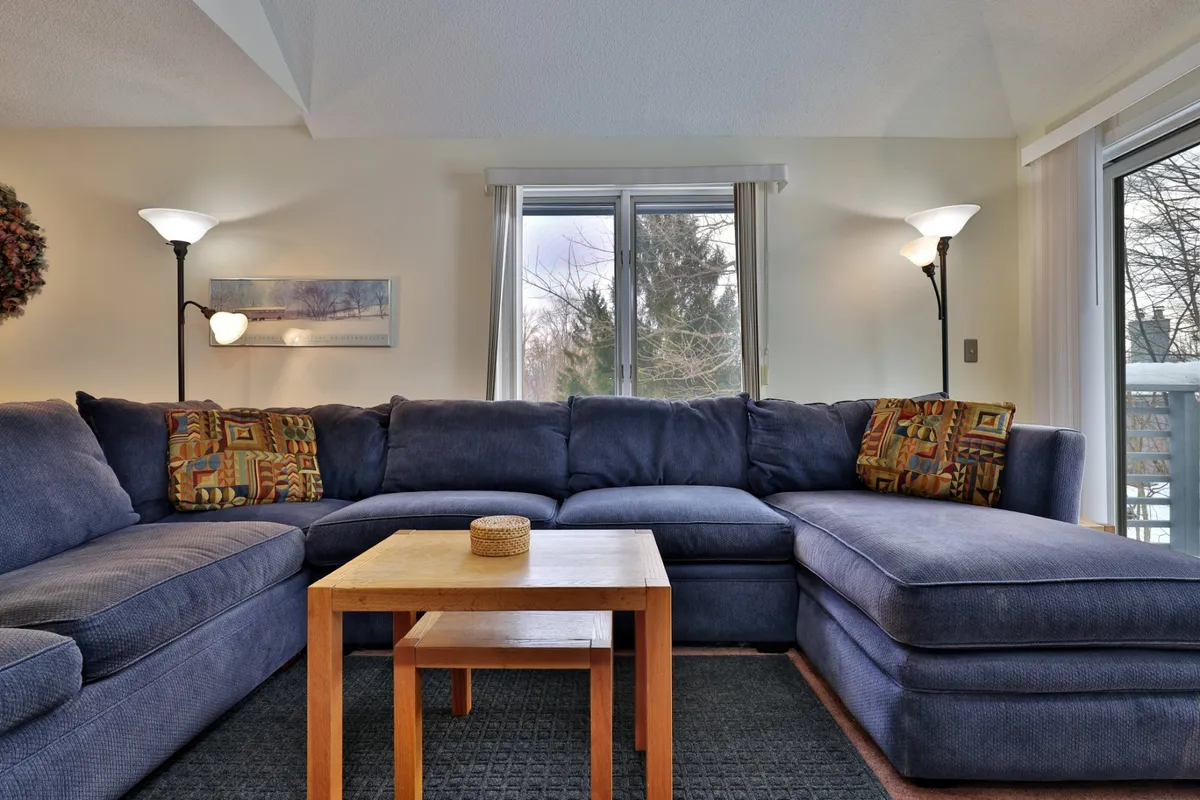 A bright, inviting living space with a blue sectional sofa, wooden coffee table, and large windows, ideal for relaxation and socializing. The decor is modern yet comforting, emphasizing both privacy and togetherness.